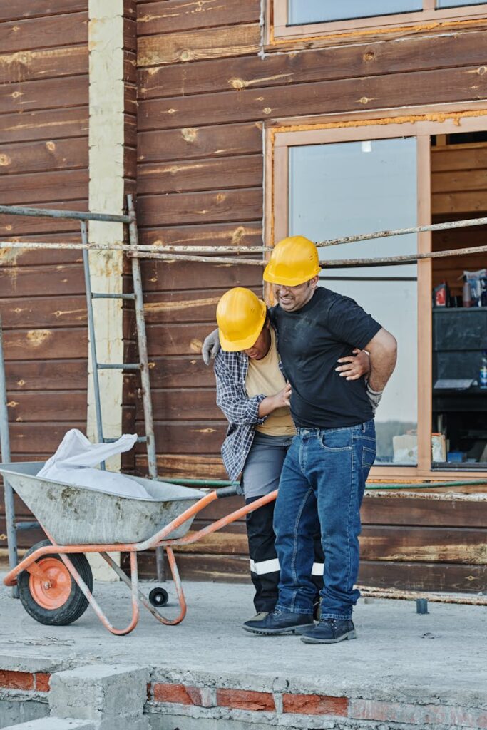 Construction workers wearing helmets assist an injured colleague at a building site with wooden cabins.